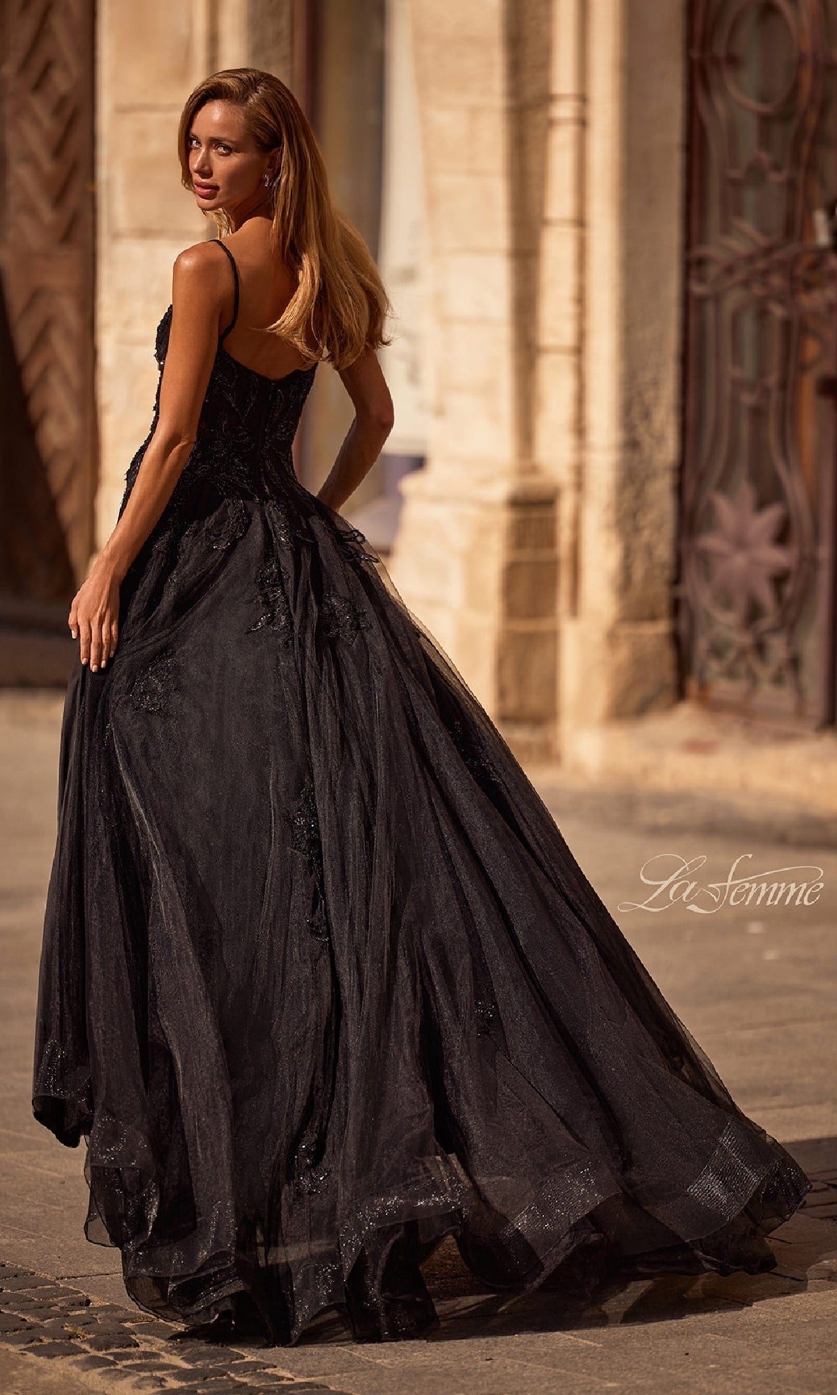 A woman with long hair walks outdoors by ornate stone buildings wearing the Long Black Glitter Prom Dress: La Femme 32781. The